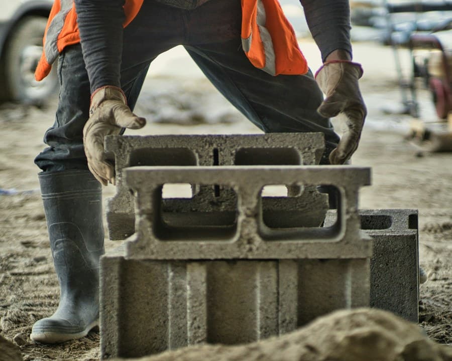 Un maçon en gilet orange soulève un parpaing sur le chantier